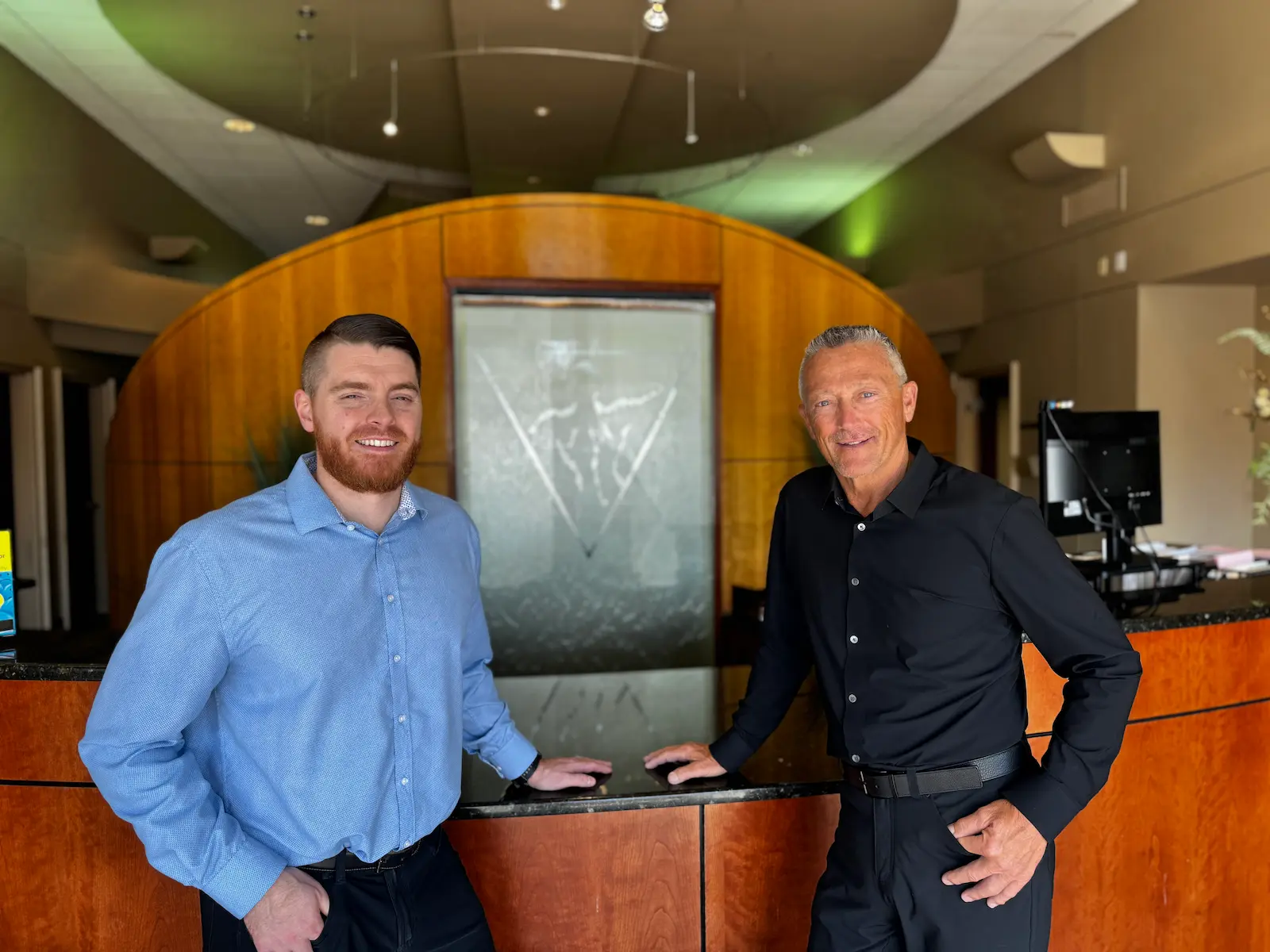 Two men, one with a red beard in a blue shirt and one with gray hair in a black shirt, stand smiling behind a reception desk with a water feature.