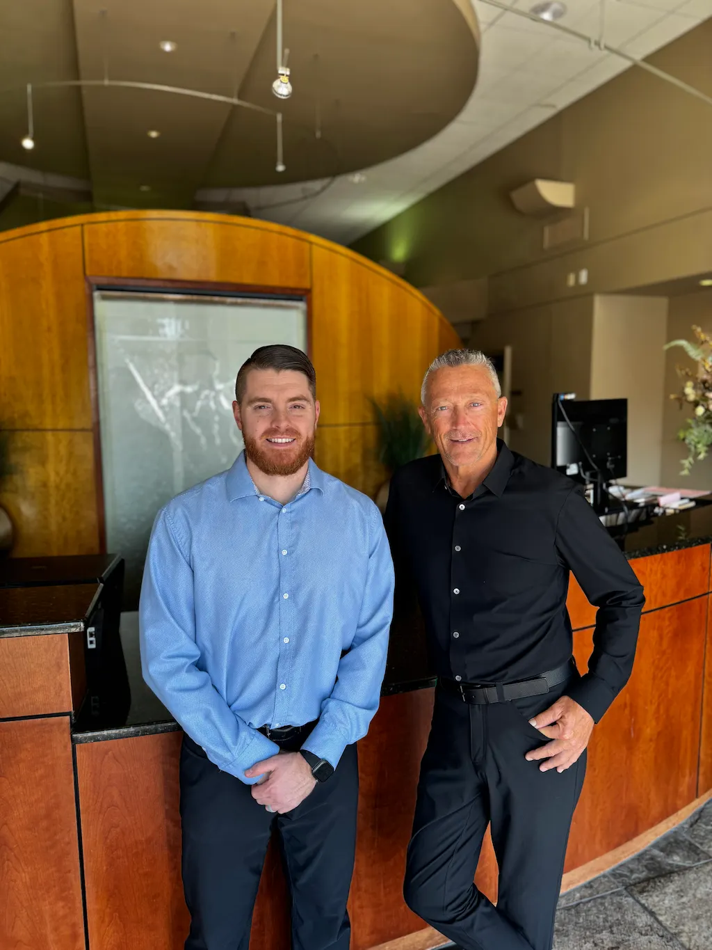 Two men, one younger with a beard in a blue shirt, and one older with gray hair in a black shirt, smiling behind a wooden reception desk.