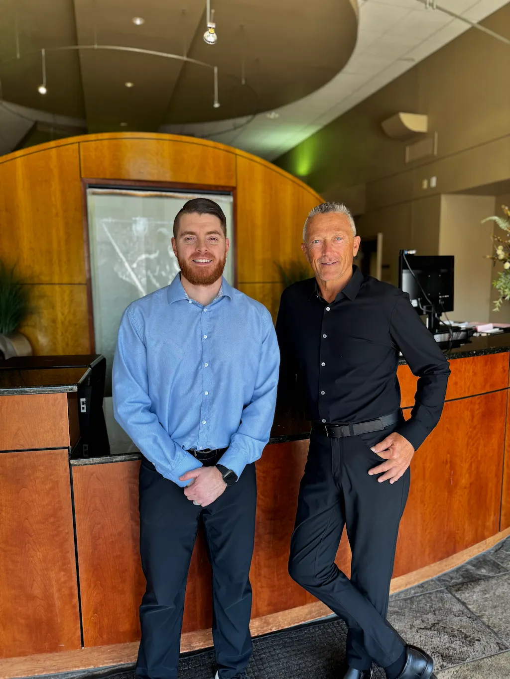 Two men, one younger with a beard in a blue shirt, and one older with gray hair in a black shirt, standing and smiling in a modern office.