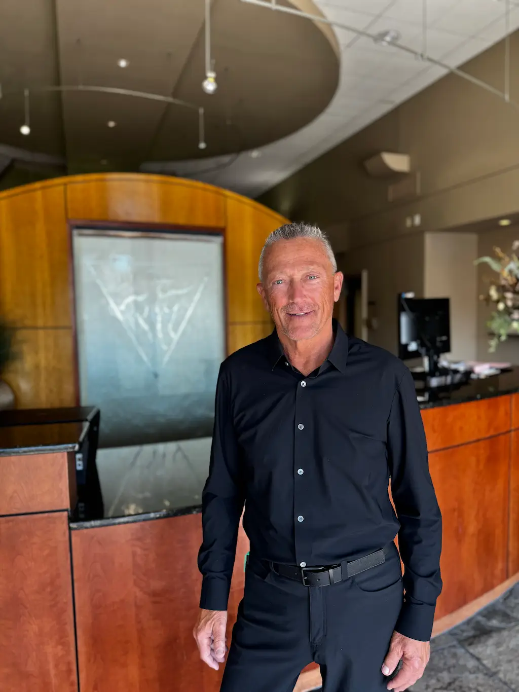 A smiling man in a black shirt stands in front of a curved wooden reception desk with a glass art panel.