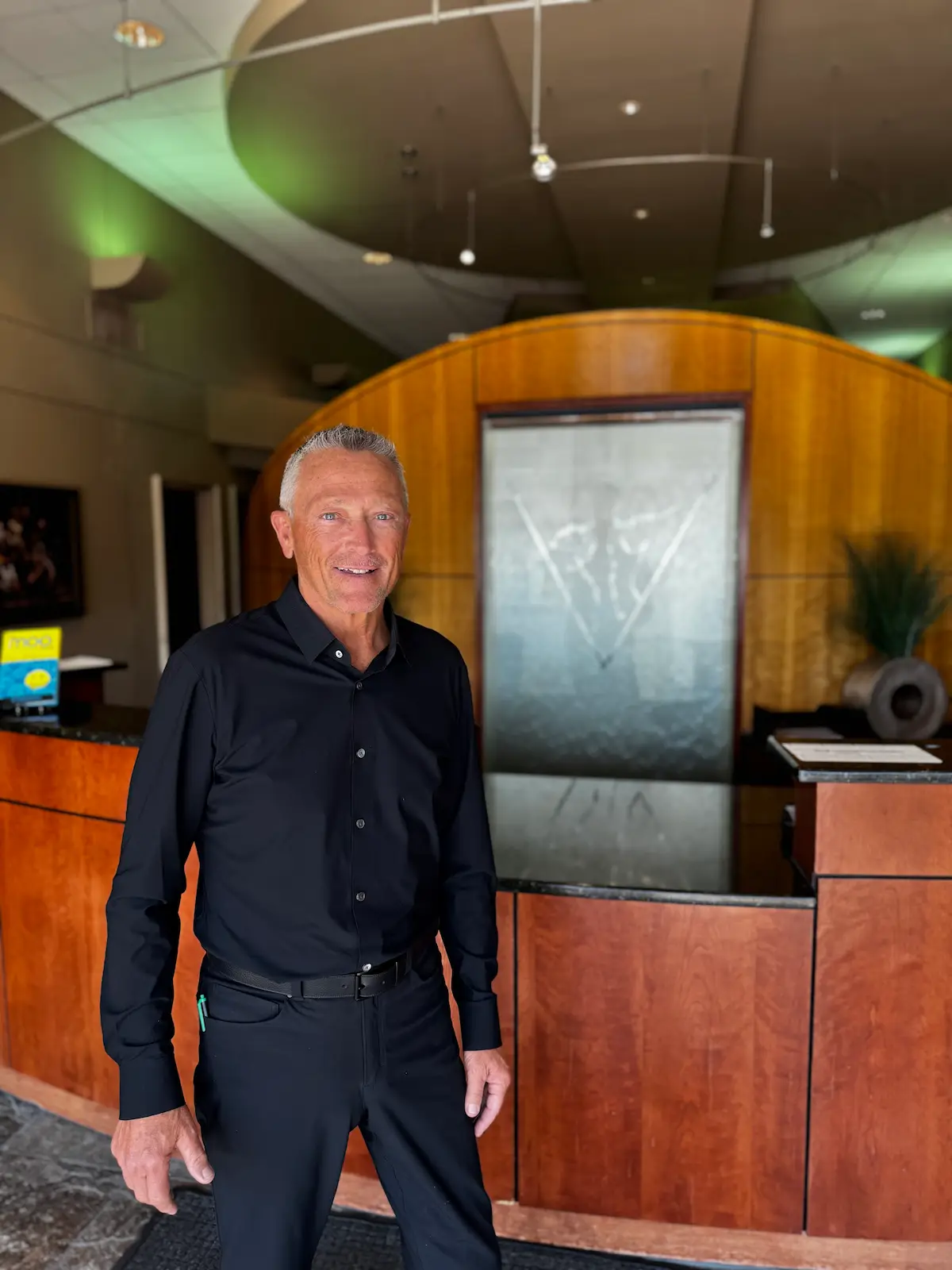 A smiling man with gray hair and blue eyes, wearing a black shirt and pants, stands in a modern reception area. Behind him is a curved wooden wall with a frosted glass panel and a dark counter.