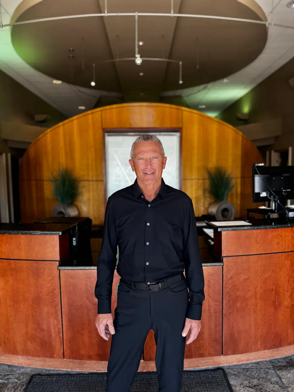 A smiling man in a black shirt and pants stands in front of a curved wooden reception desk with plants and a computer.