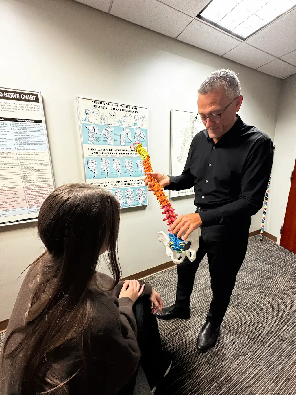 A man in a black shirt holds a colorful spine model, pointing to the pelvis, while a woman with long dark hair sits on the floor, looking at the model. Educational charts are on the wall behind them.