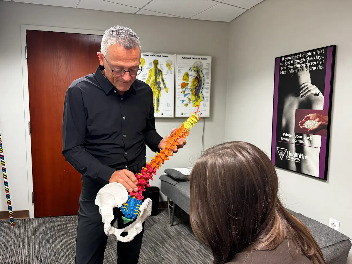 A chiropractor demonstrates a colorful spine and pelvis model to a patient in an office setting.