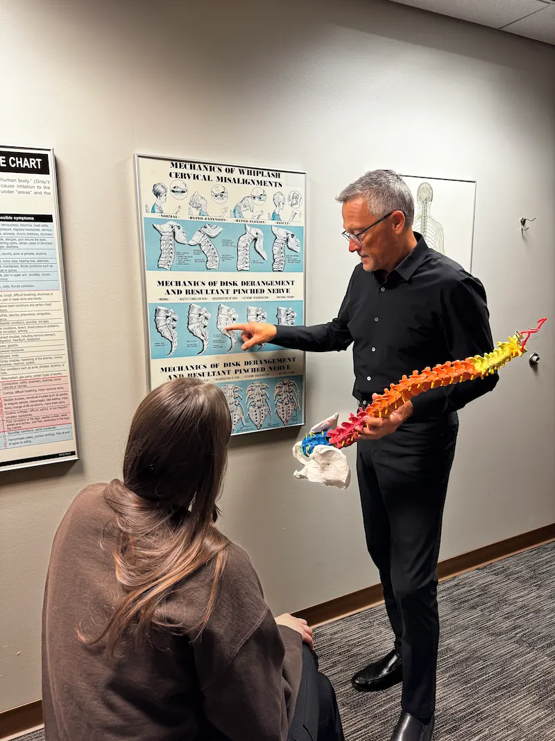 A chiropractor in a black shirt points to a spinal chart on the wall while holding a colorful spine model, explaining to a patient seated with her back to the camera.