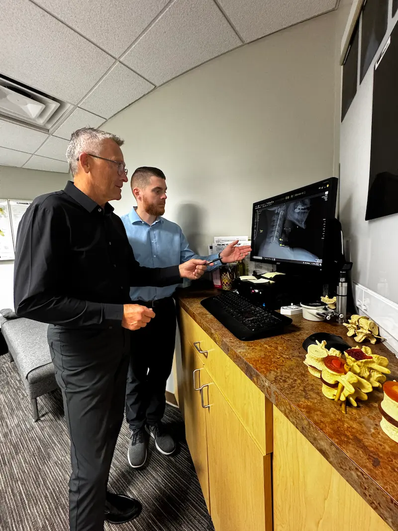Two men, one older and one younger, standing and looking at a computer screen displaying a spinal X-ray. Anatomical spine models are on the counter next to the monitor.