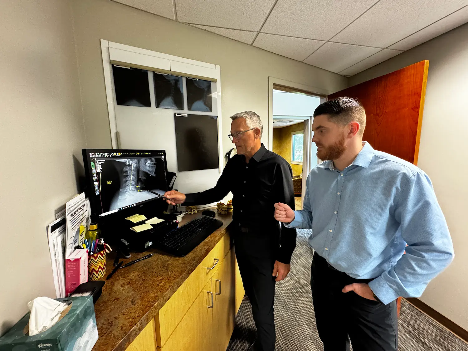 Two men, one older and one younger, stand in an office looking at a computer screen displaying spinal X-rays. The older man points at the screen.