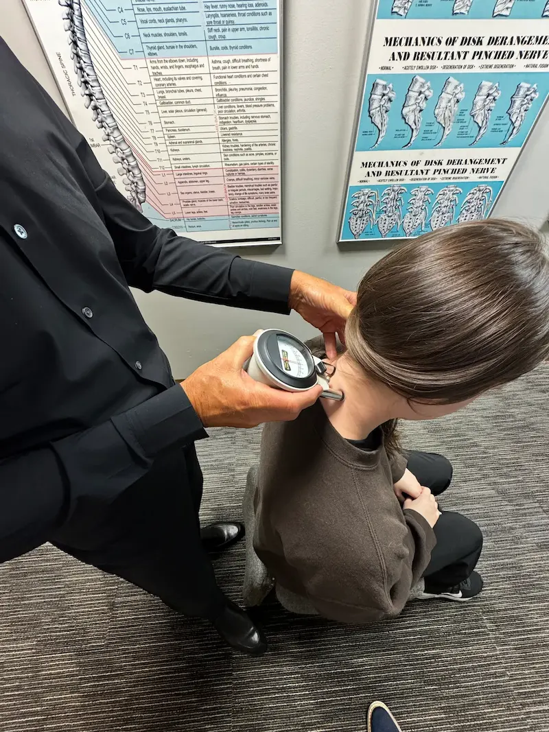A medical professional uses a handheld diagnostic device on a patient's neck, with anatomical charts visible on the wall in the background.