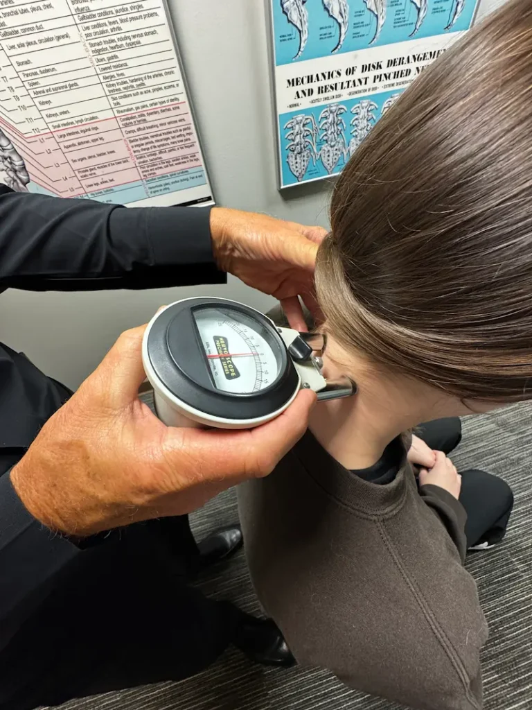 A chiropractor uses a Nervoscope on a patient's neck, with medical charts in the background.