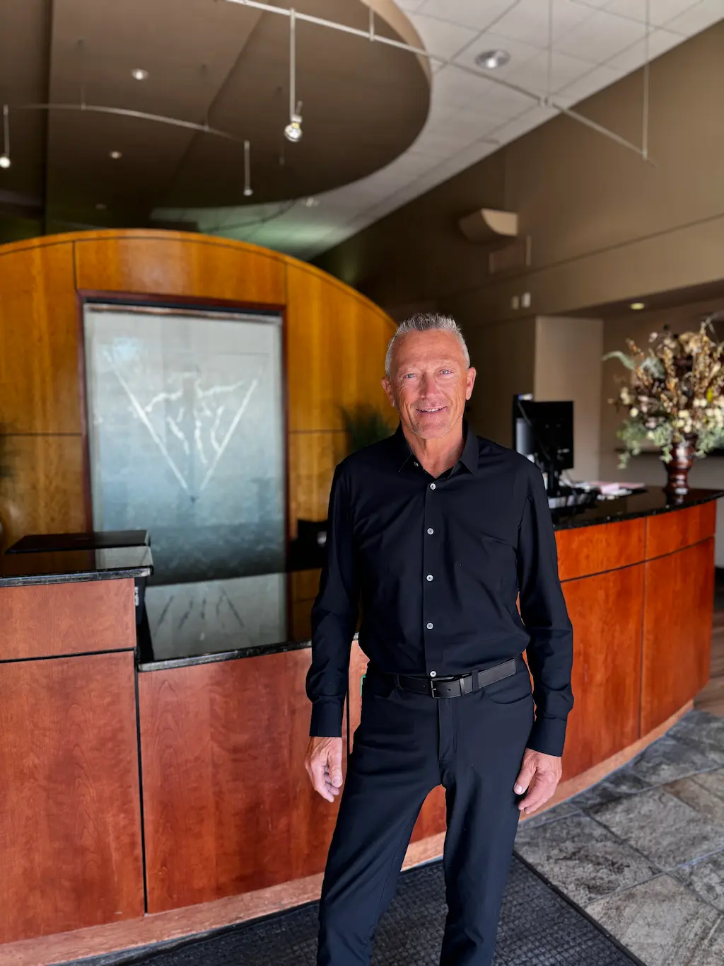 A smiling man in a black shirt and pants stands in a modern reception area with a wooden desk and a water feature.