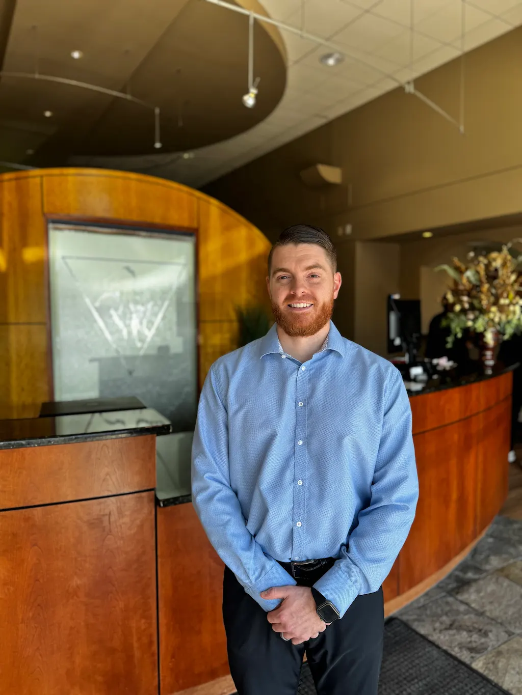 A smiling man with a red beard in a blue shirt stands in a lobby with a wooden reception desk and floral arrangement.