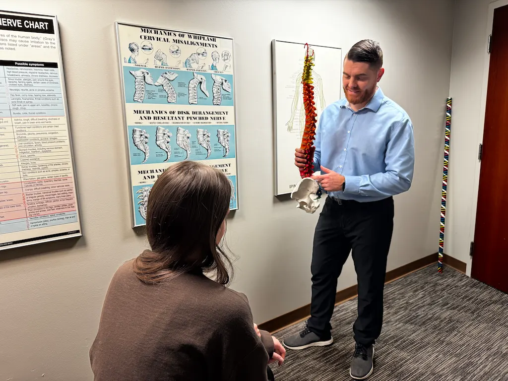 A man holding a spine and pelvis model explains to a woman in an office with anatomical charts on the wall.