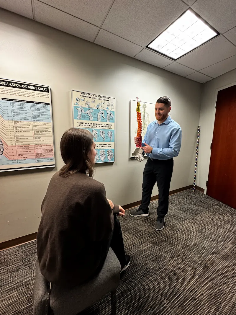 A male chiropractor explains a colorful spine model to a female patient in a consultation room with anatomical charts on the wall.