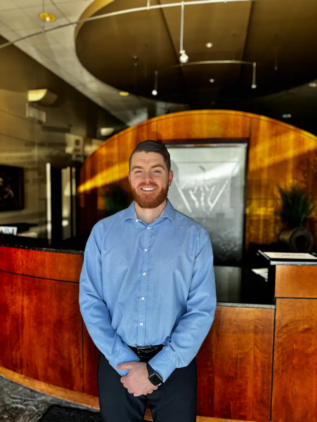 A smiling man with a beard, wearing a blue shirt and black pants, stands in front of a curved wooden reception desk with a water feature behind him.