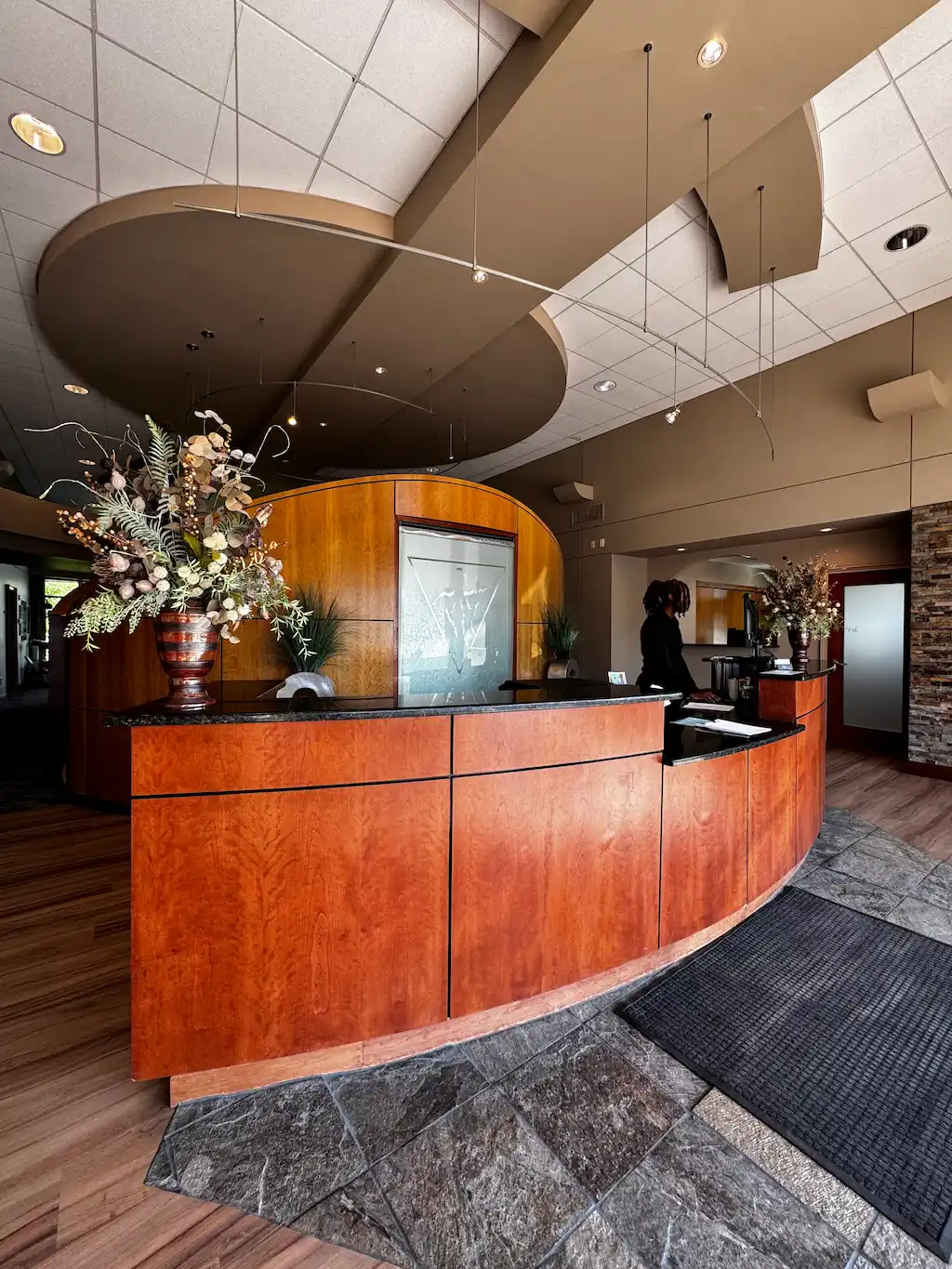 A modern reception area with a curved wooden desk, black countertop, and large floral arrangements. A person stands behind the desk. The ceiling has curved brown bulkheads and track lighting. The floor is a mix of wood and stone tiles.