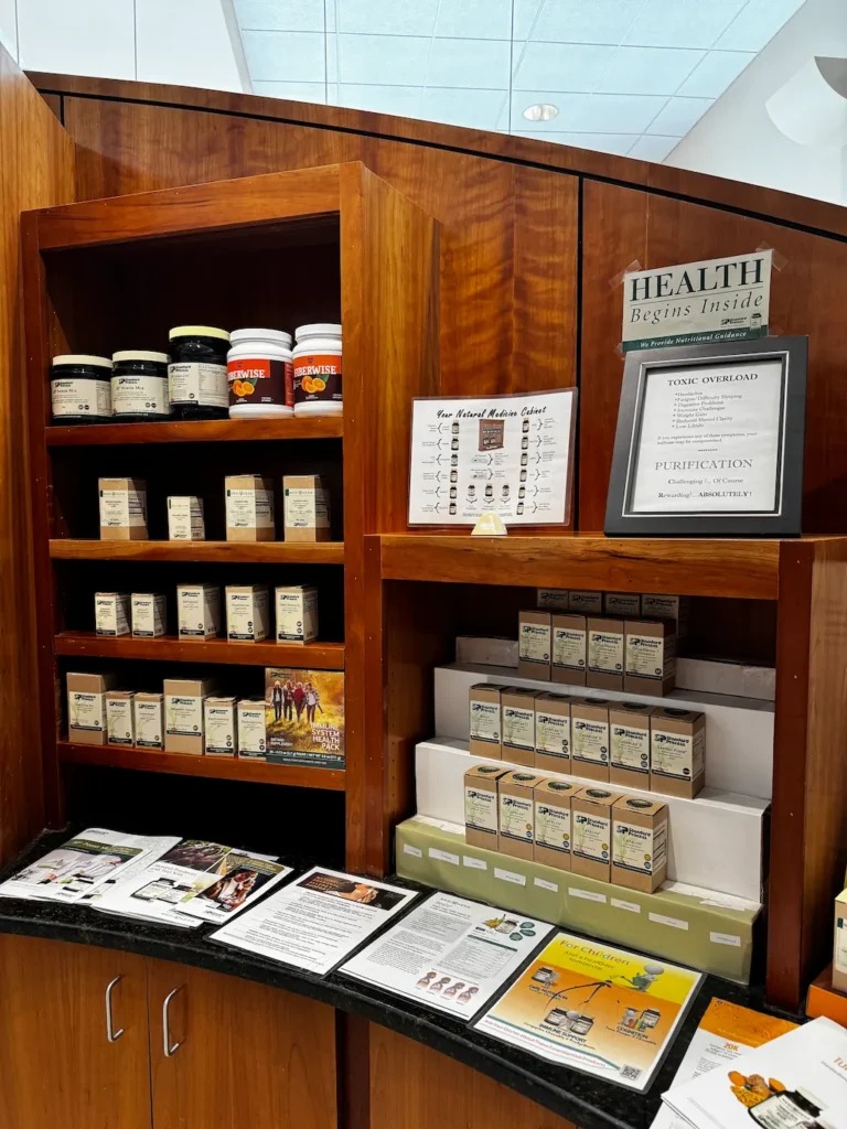 A wooden display cabinet showcasing various health supplements like Fiberwise and Standard Process products, alongside informational signs about 'Toxic Overload' and 'Purification,' and brochures for children's health.