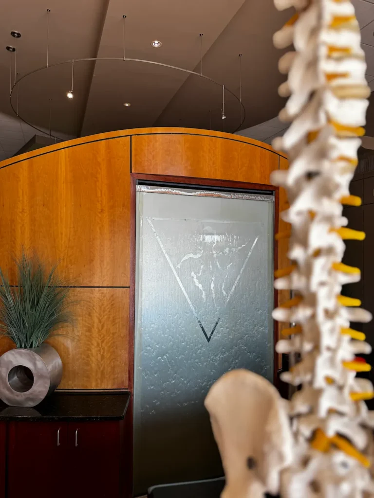 A blurred anatomical spine model in the foreground, with a curved wooden wall and a glass water feature with a triangular design in the midground. A modern planter with artificial grass is on a counter. The ceiling has circular track lighting.