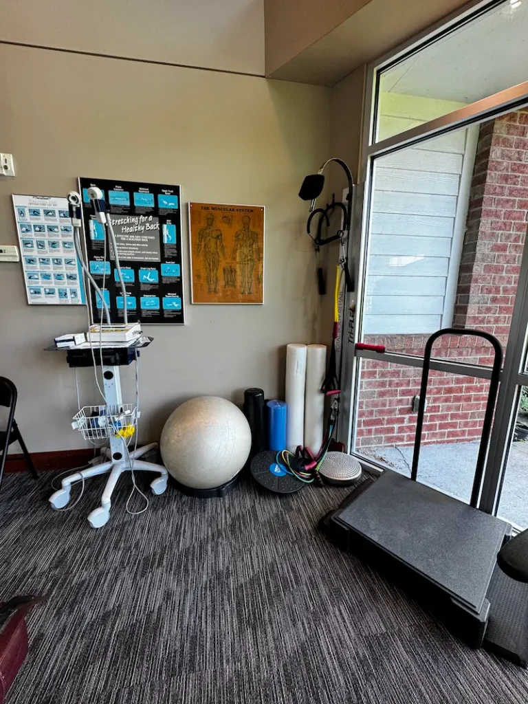 A physical therapy room with exercise equipment, including an exercise ball, foam rollers, resistance bands, and anatomical charts on the wall.
