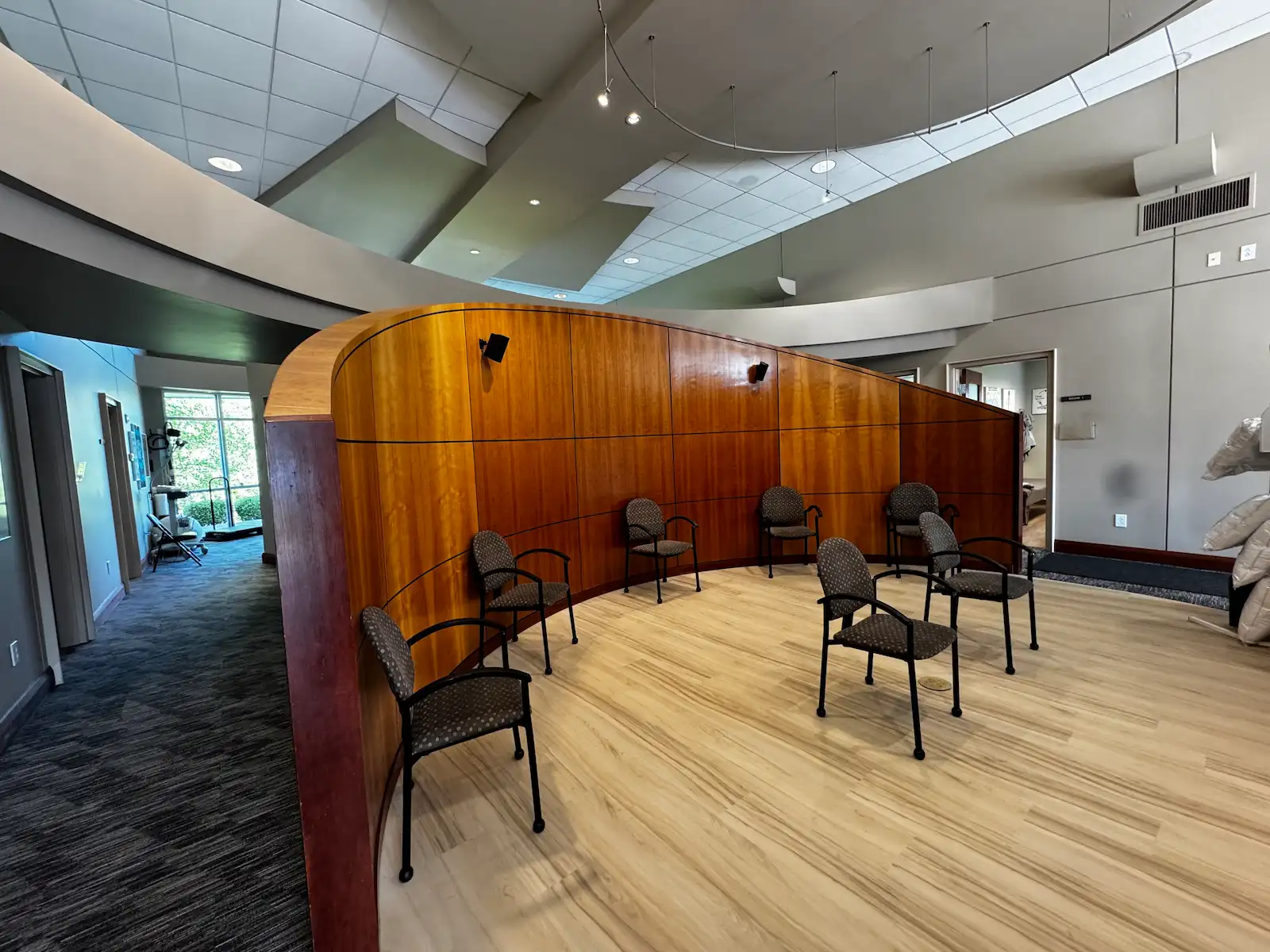 A waiting room with a curved wooden wall, light wood floor, and multiple chairs arranged along the wall.