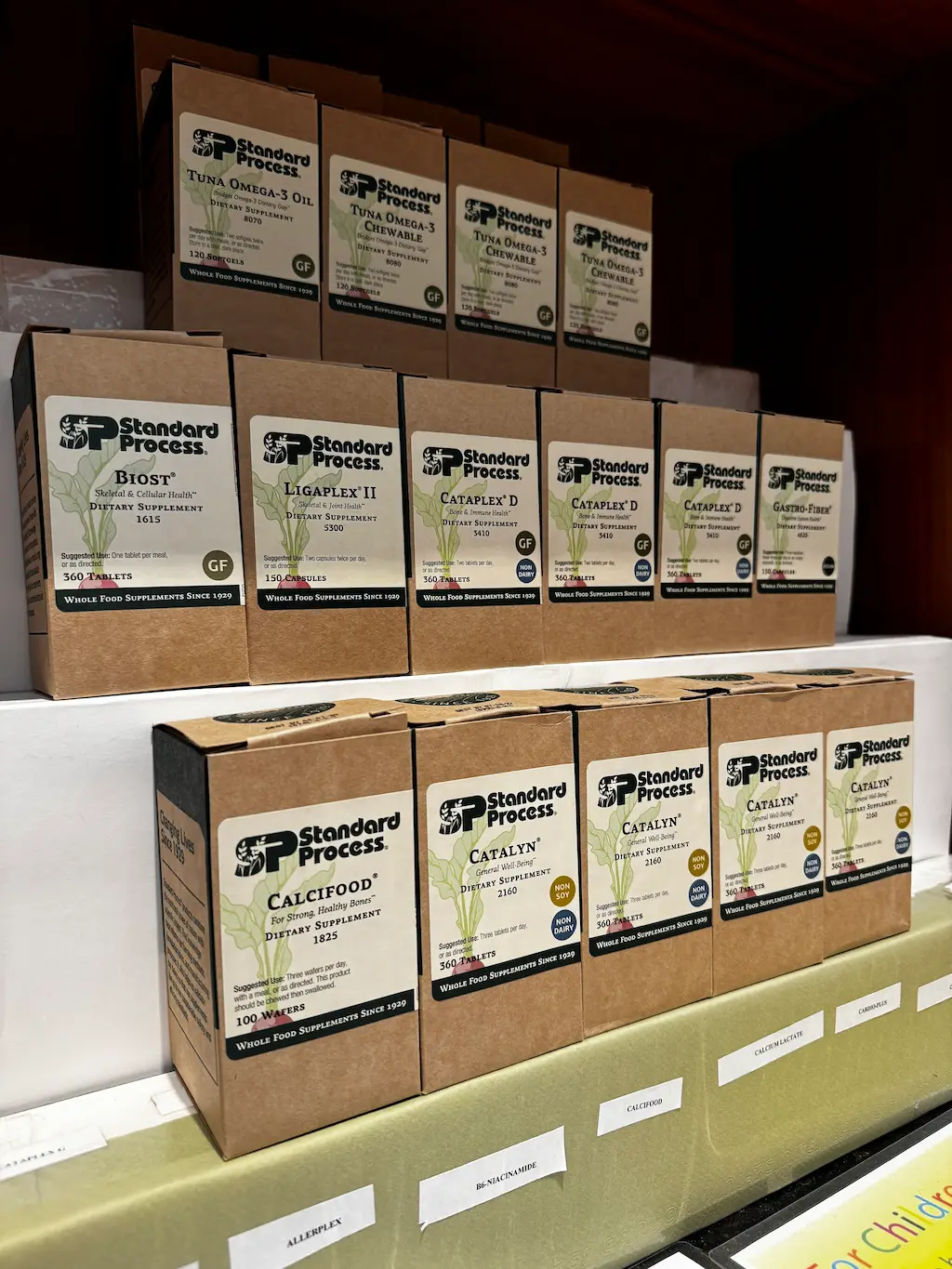 Shelves filled with various Standard Process dietary supplements in brown and white packaging, including Tuna Omega-3, Biost, Cataplex D, Calcifood, and Catalyn.