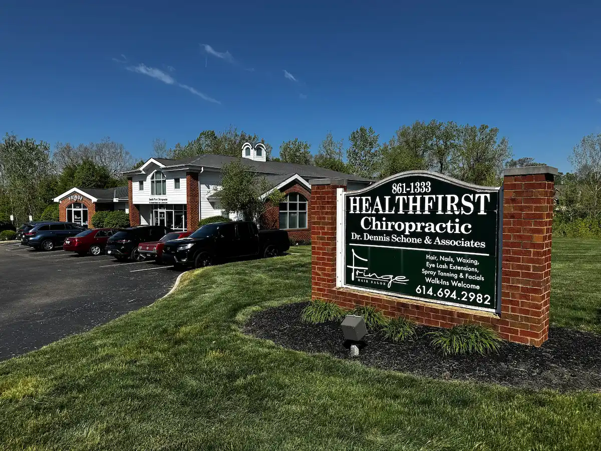 Exterior view of a commercial building with a brick and white facade, housing HealthFirst Chiropractic and Fringe Hair Salon. A large sign in the foreground lists both businesses and their services, with a parking lot full of cars and green landscaping under a blue sky.