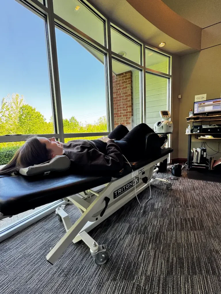 A woman lies on her back on a black medical table in a room with large windows overlooking trees and buildings.