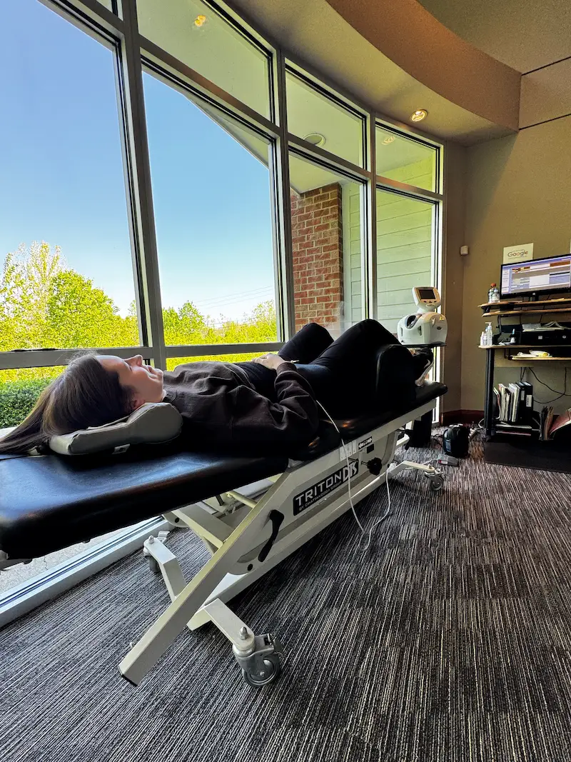 A woman lies on her back on a black medical table in a room with large windows overlooking trees and buildings.