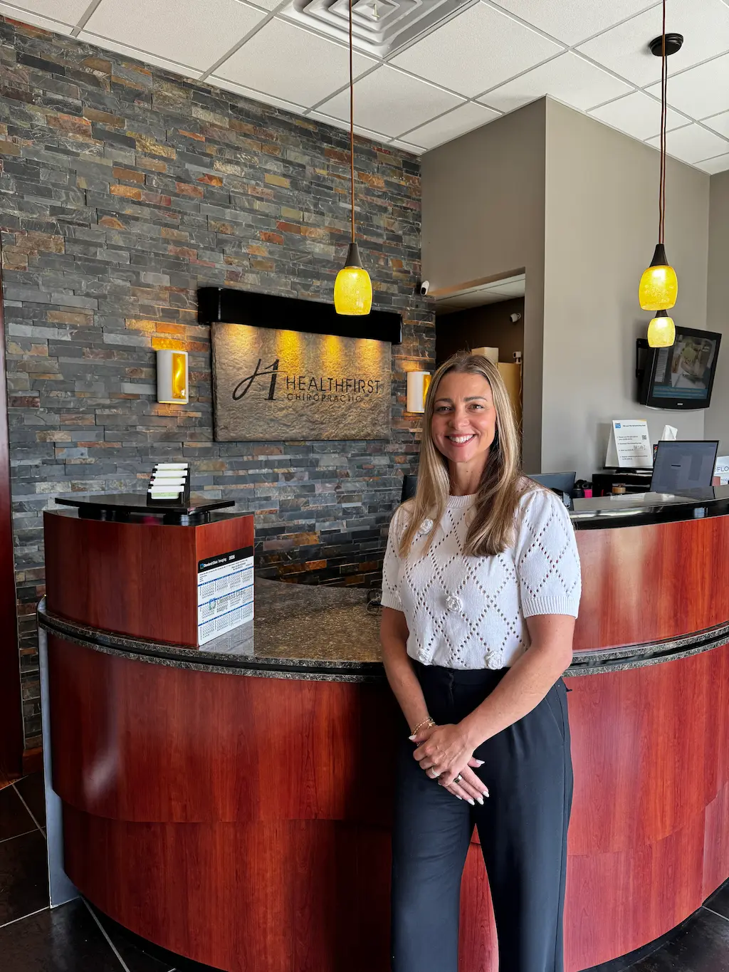 A smiling woman stands in front of a curved reception desk and a stone wall with a 'HealthFirst Chiropractic' sign.