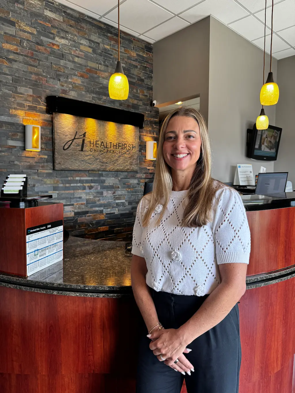 A smiling woman with long brown hair stands in front of a reception desk at HealthFirst Chiropractic, with a stone wall and company sign behind her.