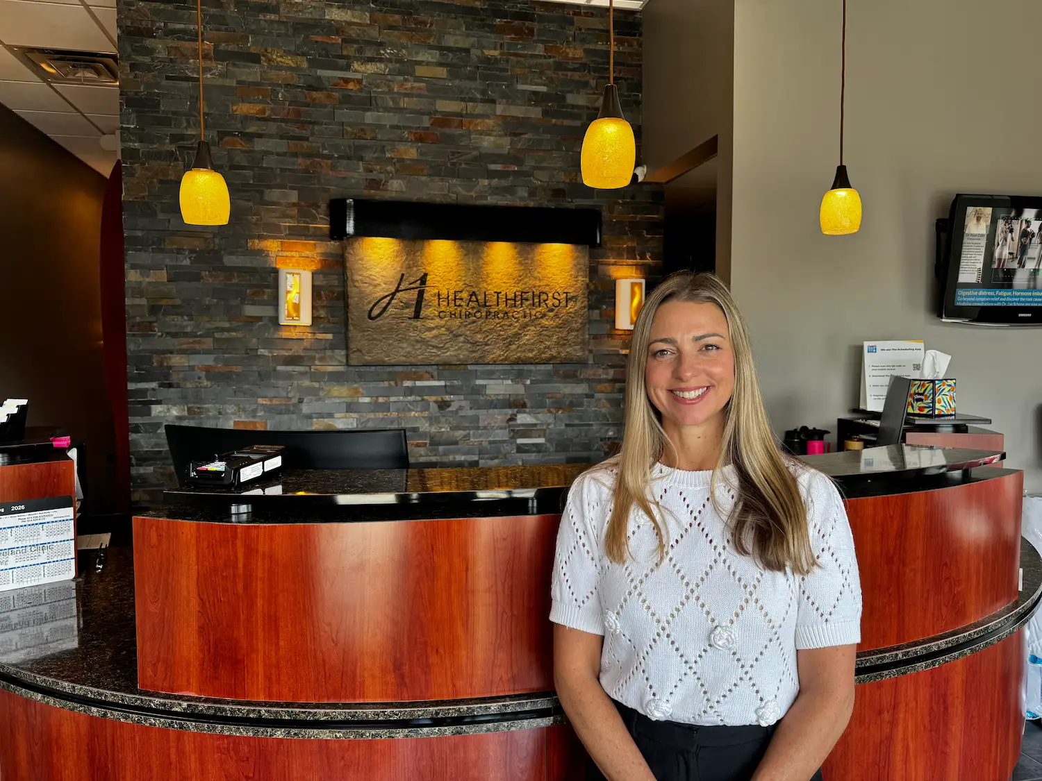 A smiling woman stands in a chiropractic office reception area with a stone wall and 'HealthFirst Chiropractic' sign behind her.