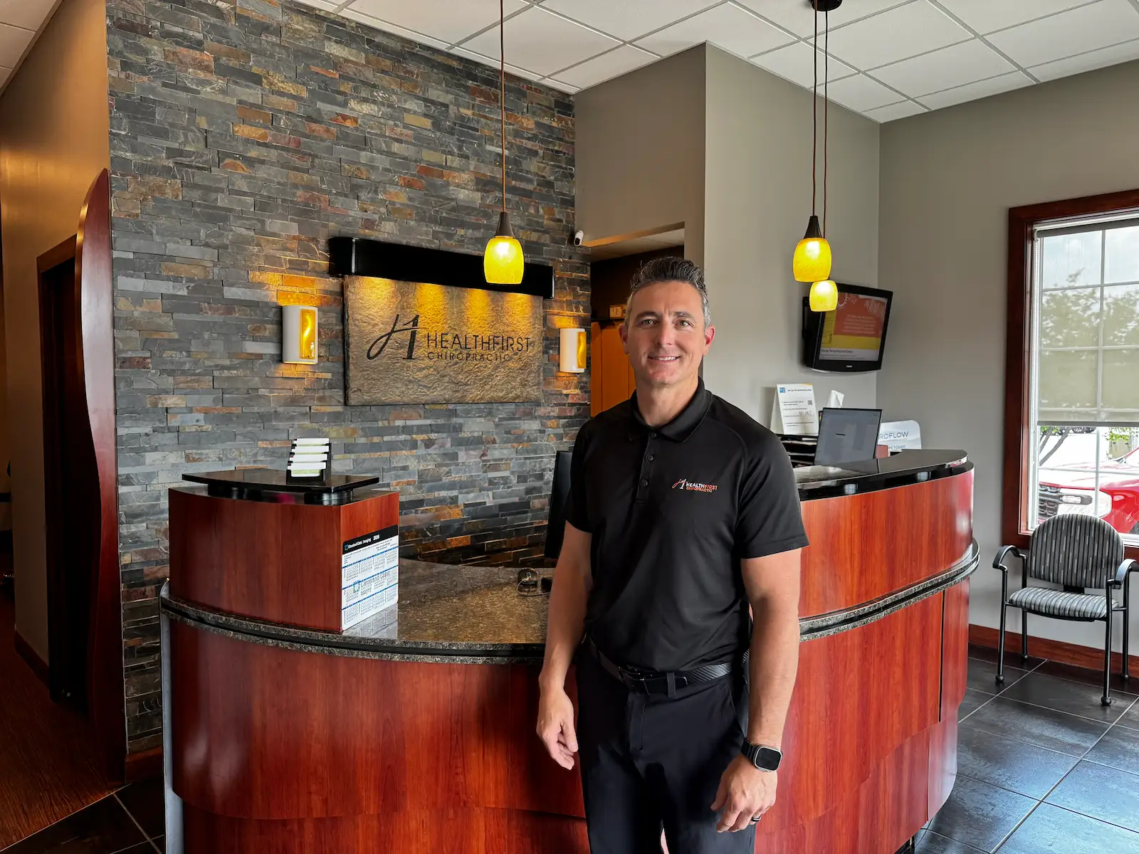 A smiling man in a black polo shirt stands in the reception area of HealthFirst Chiropractic, with a stone-tiled wall and reception desk behind him.