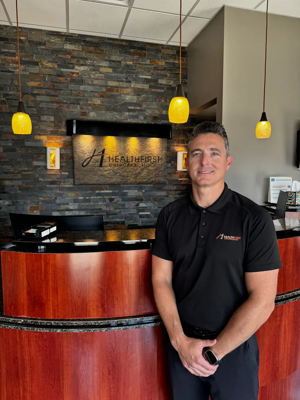 A smiling man in a black polo shirt stands in front of a reception desk with a 'HealthFirst Chiropractic' logo on a stone wall behind him.