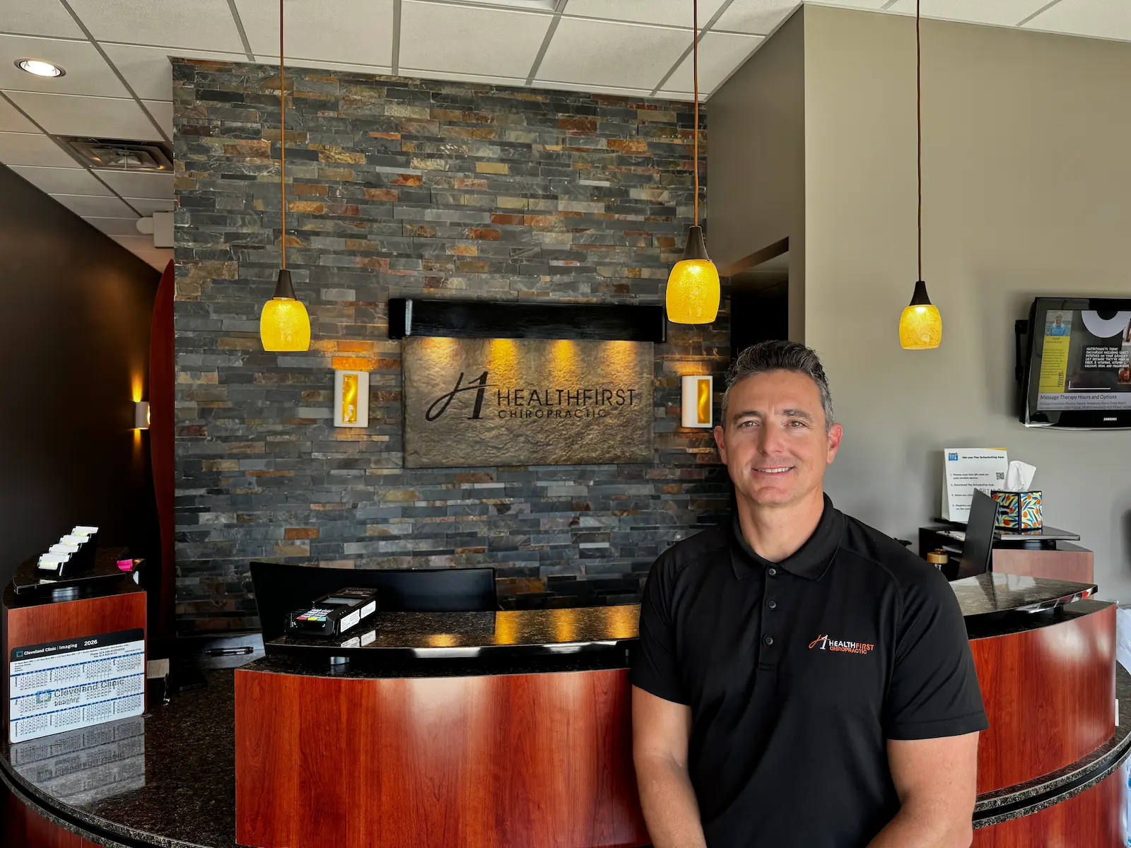 A man in a black polo shirt with a 'HealthFirst Chiropractic' logo stands smiling in front of a reception desk and a stone wall with a backlit 'HealthFirst Chiropractic' sign.