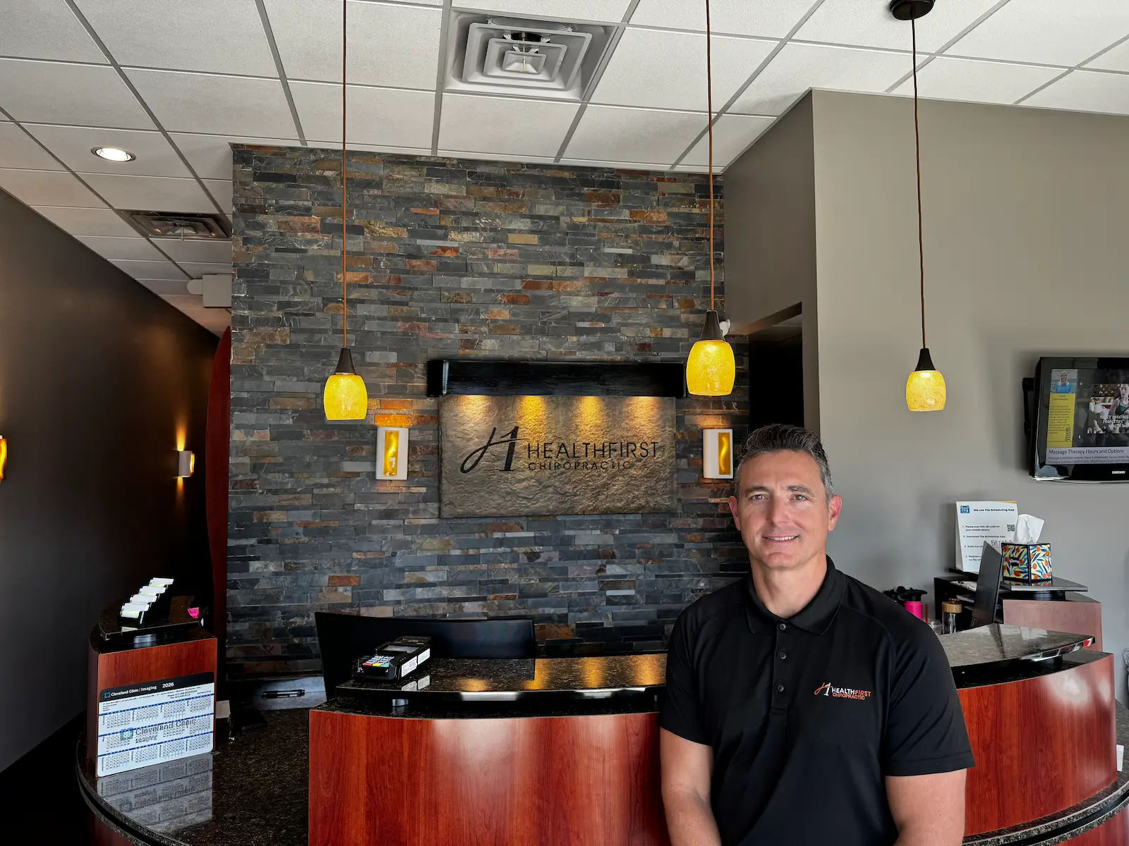 A smiling man in a black polo shirt stands in a modern chiropractic office reception area with a stone-tiled wall and a 'HealthFirst Chiropractic' sign.