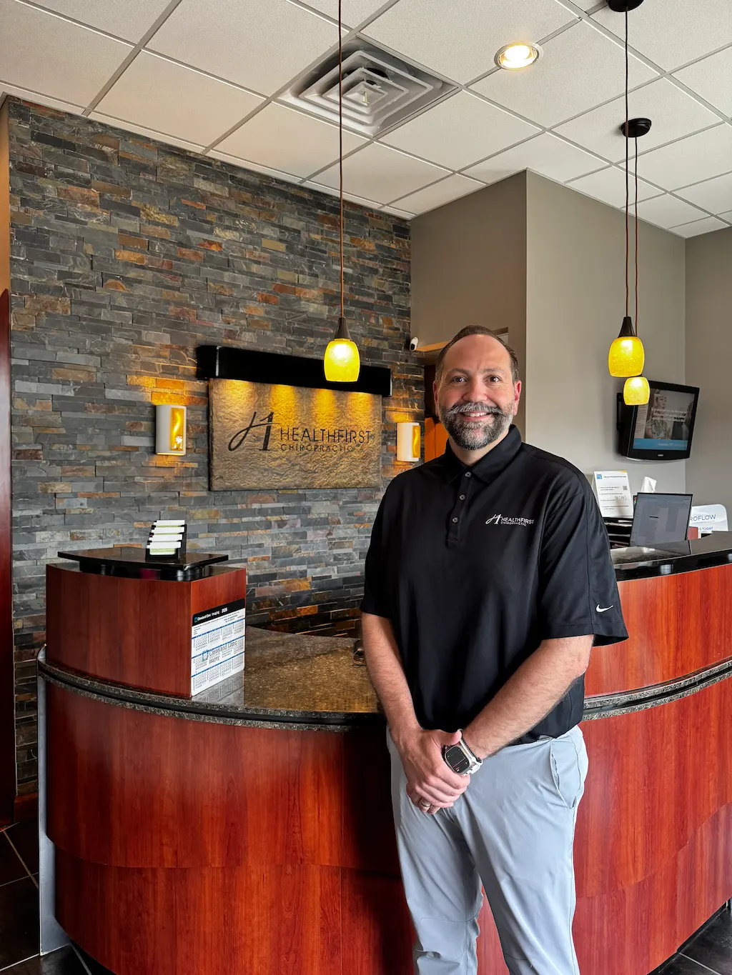 A smiling man in a black polo shirt stands in a modern chiropractic office reception area with a 'HealthFirst Chiropractic' sign on a stone wall behind him.