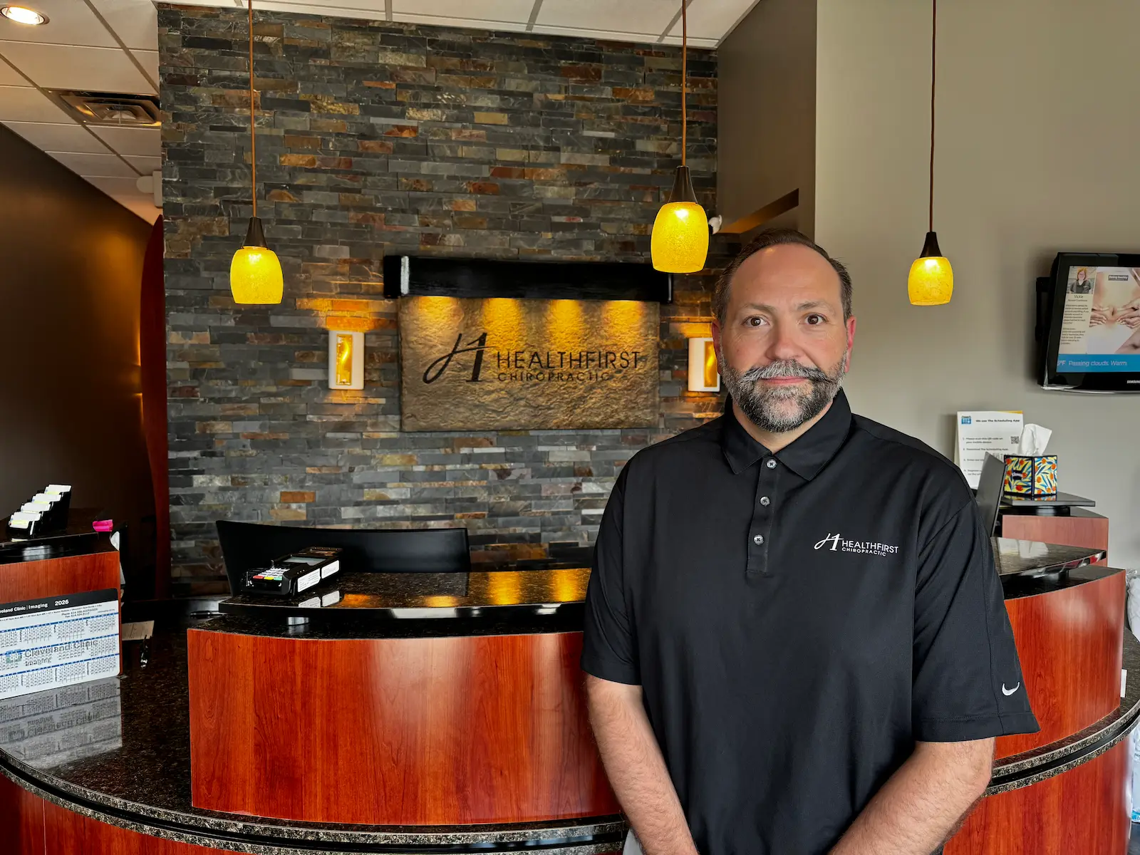 A man in a black polo shirt with a company logo stands in a chiropractic office reception area.