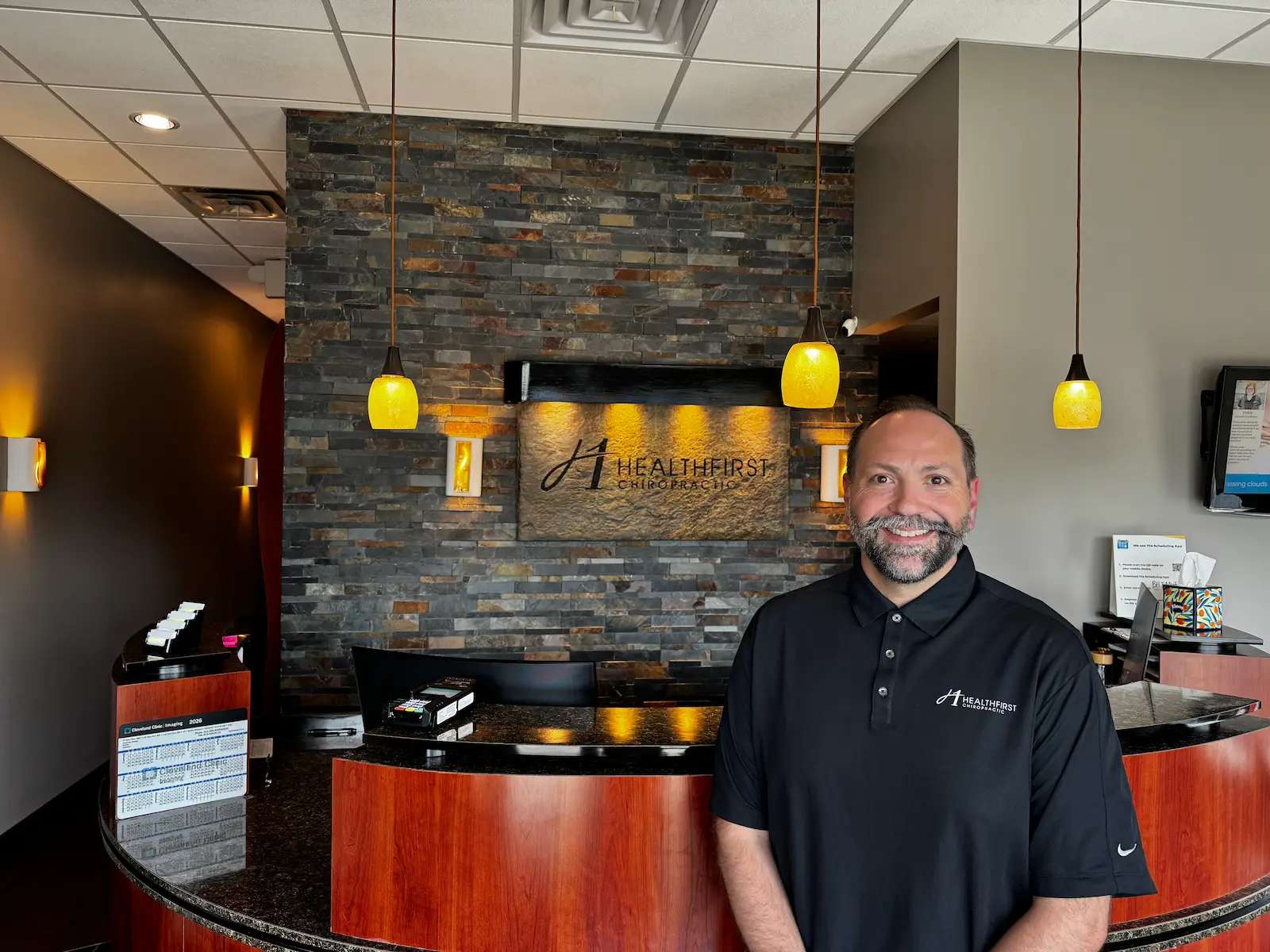 A smiling man in a black polo shirt stands in a modern chiropractic office reception area with a stone wall and 'HealthFirst Chiropractic' sign behind him.