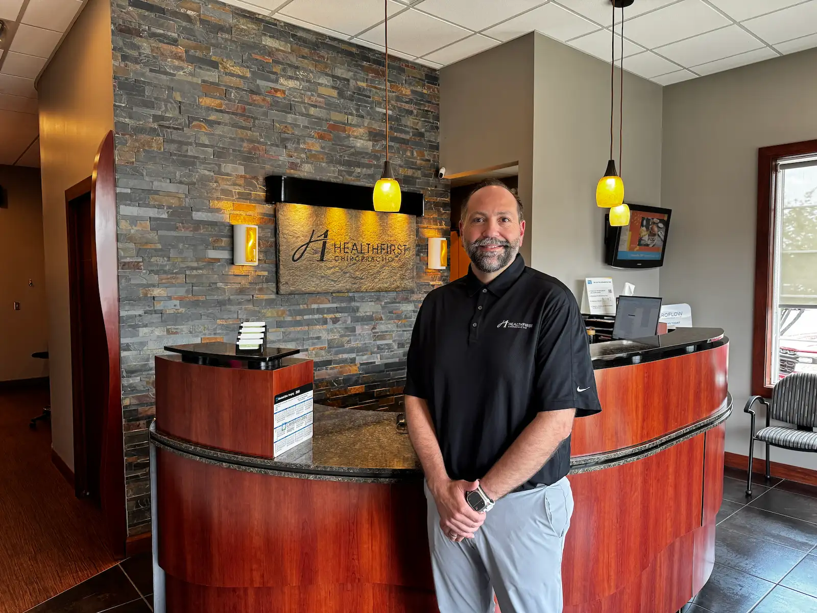 A smiling man in a black polo shirt stands in front of a curved wooden reception desk with a stone-tiled wall behind him, featuring a "HealthFirst Chiropractic" sign.