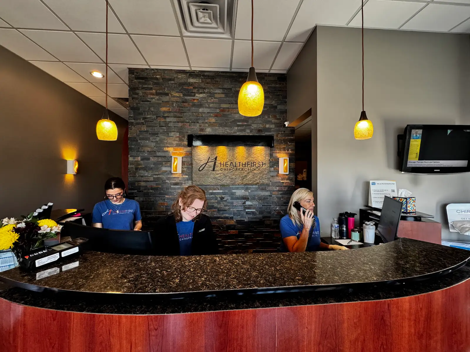 Three women in blue Healthfirst Chiropractic shirts work at a curved reception desk in a chiropractic office. A stone-tiled wall with the company logo is behind them.