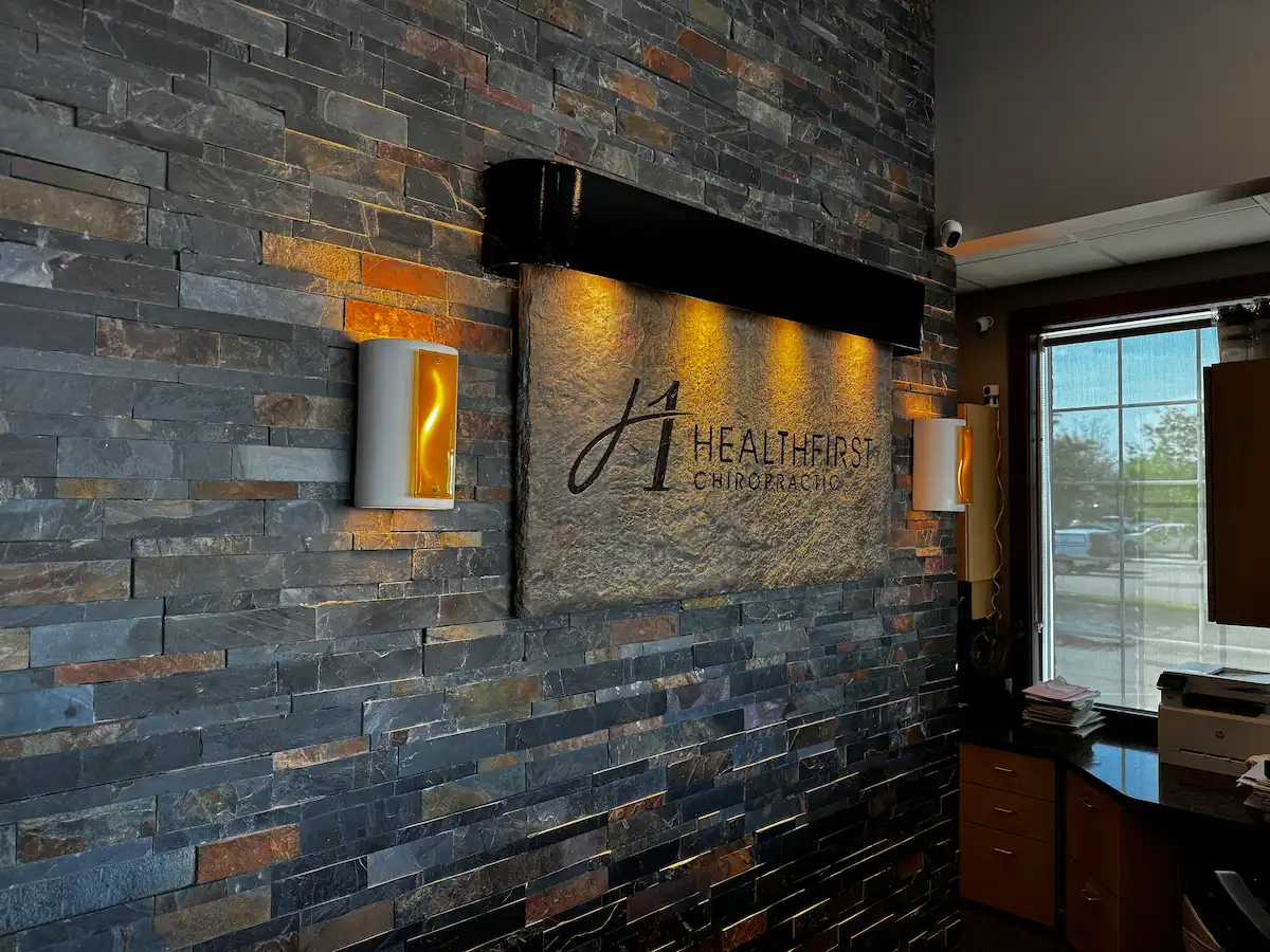 Reception area with a stone wall featuring the 'HealthFirst Chiropractic' sign illuminated by warm spotlights and sconces. A window and reception desk are visible on the right.
