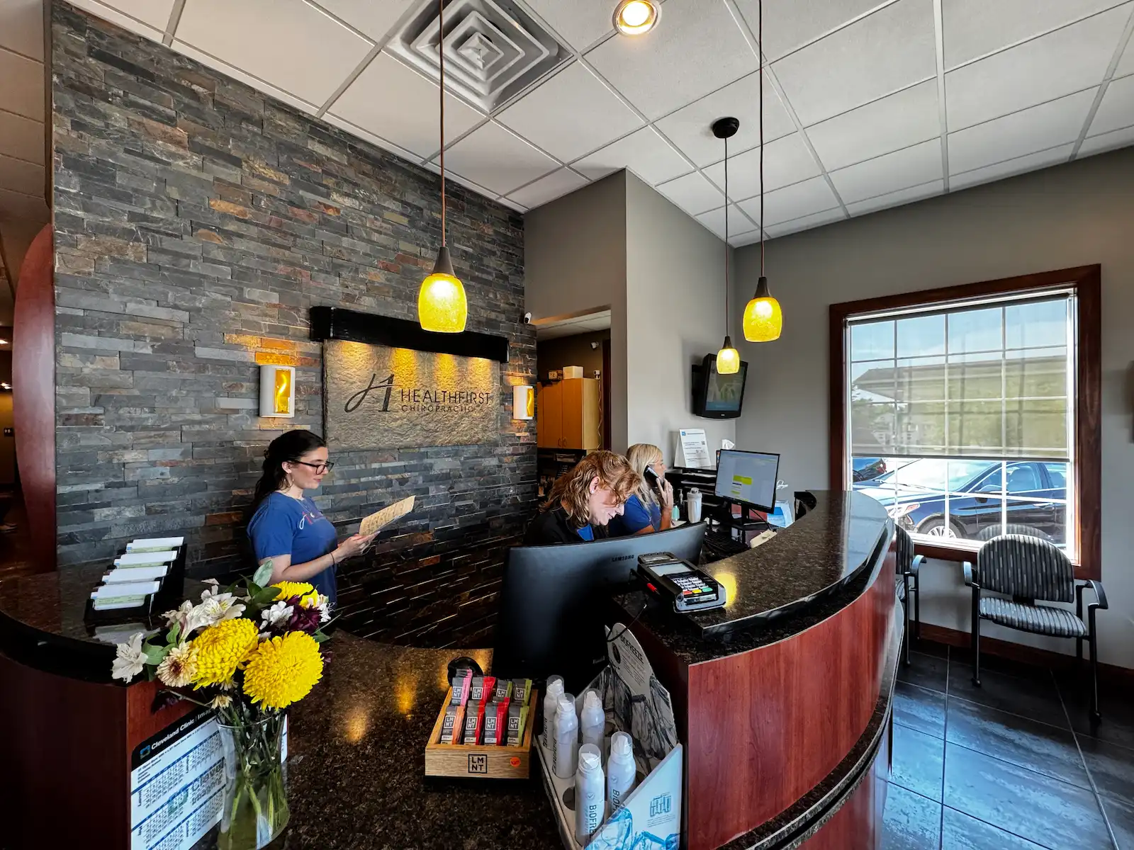 A modern chiropractic reception area with a curved desk, three staff members, a stone accent wall with the 'Healthfirst Chiropractic' logo, and yellow pendant lights.