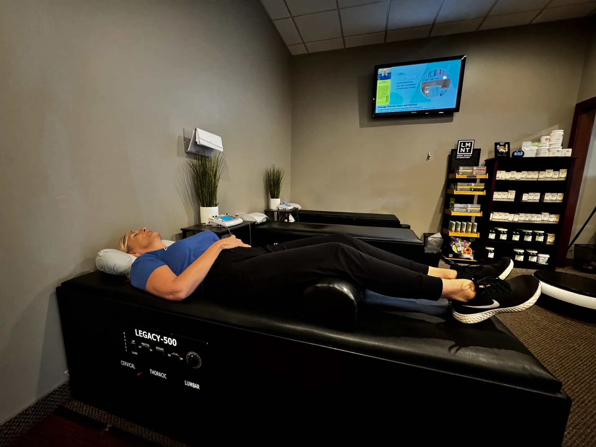 A woman lies on her back on a black Legacy-500 therapy table in a wellness center, with a TV and product shelves in the background.