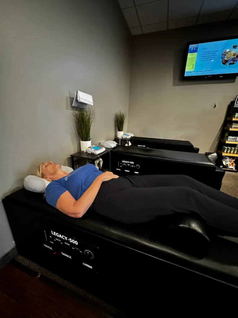 A woman lies on her back on a black LEGACY-500 therapy table with a neck pillow and knee support in a modern clinic setting.