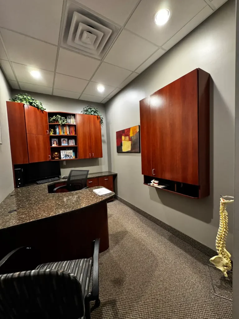 An L-shaped office desk with a granite countertop, dark wood cabinets, a computer, and a human spine model in a professional room.