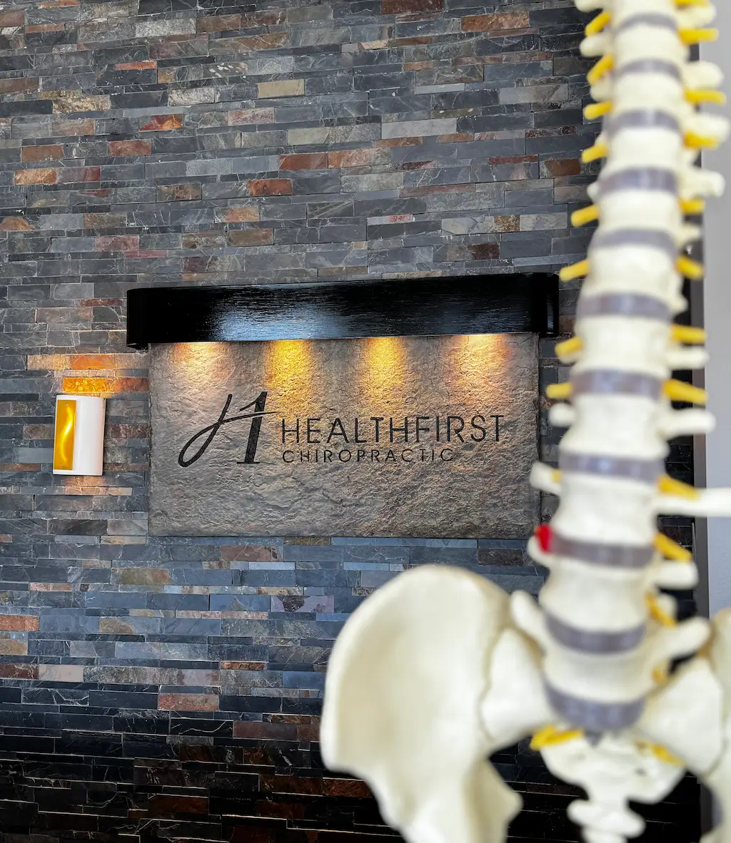 A Healthfirst Chiropractic sign on a dark, multi-toned stone wall, illuminated by spotlights. A blurred anatomical model of a human spine and pelvis is in the foreground.