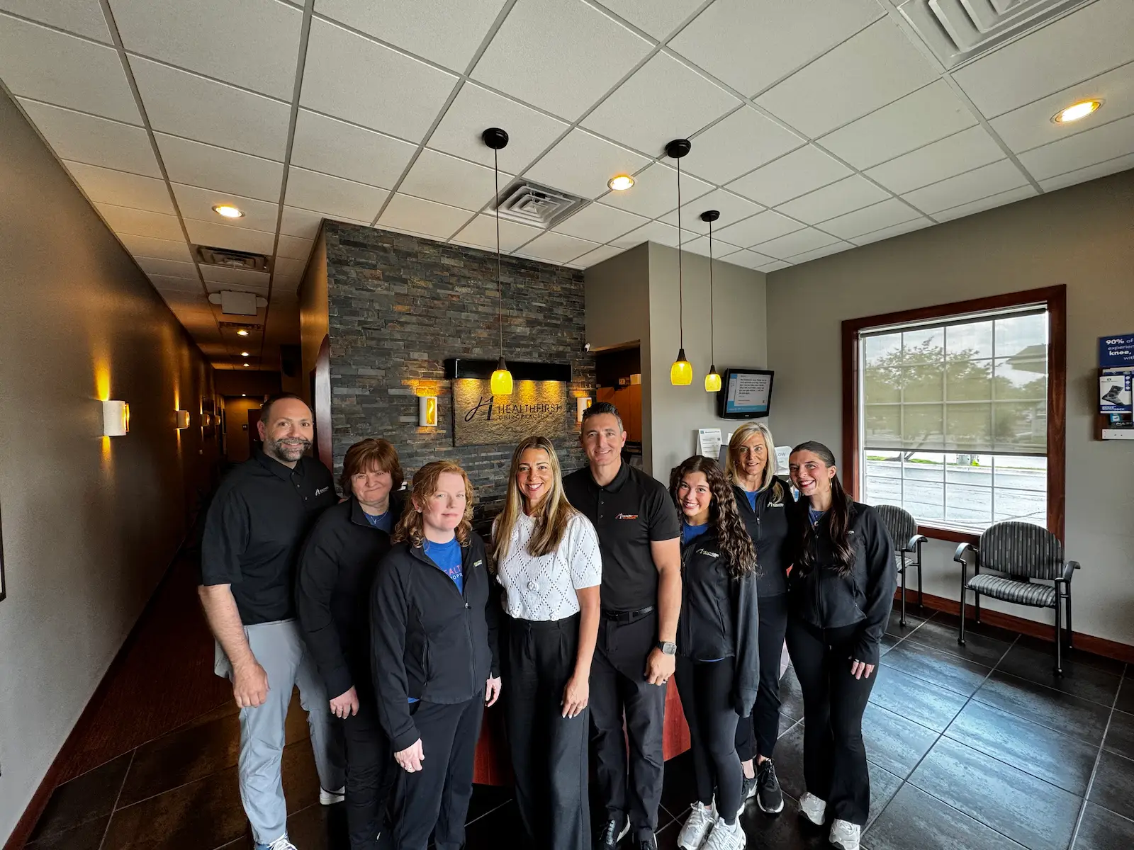 A group photo of nine smiling staff members, five women and four men, in the reception area of Healthfirst, with a stone-textured wall and logo behind them.
