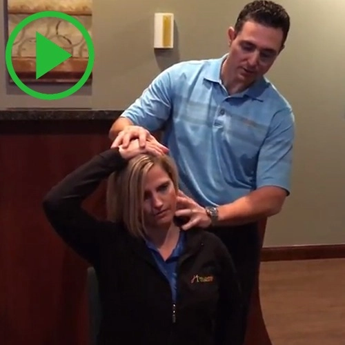 A man in a blue shirt stands behind a seated woman, holding her head and neck. A green play button is overlaid on the image.