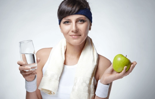 A woman in athletic wear holds a glass of water in one hand and a green apple in the other, smiling at the camera.