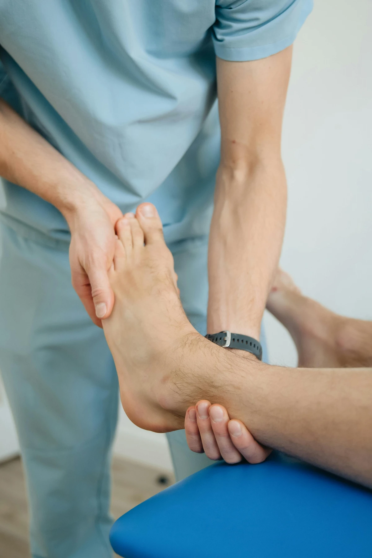 Close-up of a therapist's hands in light blue scrubs holding and examining a patient's bare foot, which rests on a blue table.