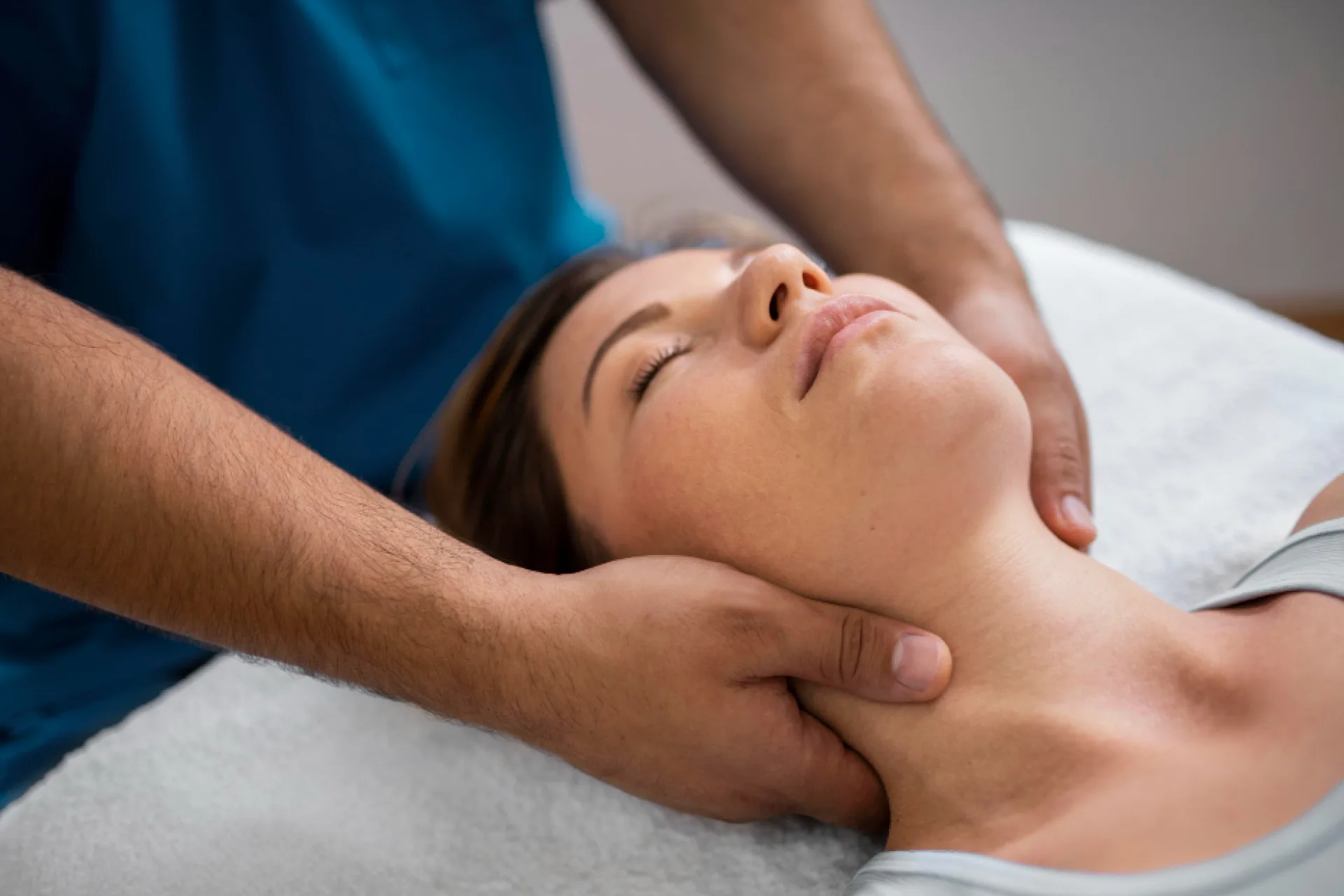Close-up of a woman lying down with her eyes closed, receiving a neck massage from a therapist whose hands are on her neck.